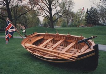 small wooden boat lying slightly tilted on green grass and flying Union Jack flag. It has four sets of oarlocks and four thwarts, two with holes for sail masts.