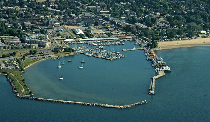 a colour aerial photograph of Cobourg Harbour showing a long breakwater at the bottom almost meeting the end of another pier to the right, leaving a narrow entrance into the harbour. The water is blue and inside the harbour sailboats are anchored or moored at a marina. On the shore from left to right are condominium buildings large tents erected for the Cobourg Festival and trailers parked in front of a sandy beach. In the distance are business buildings, residences and green trees.