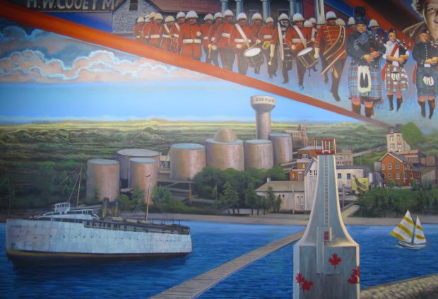 a colourful mural showing the current white Cobourg lighthouse to the right and a large white ferry to the left. On shore, in the distance to the left and centre, are seven large silver oil tanks and a water tower, and to the right beyond the lighthouse are a few businesses . Above them are depicted the red uniformed Cobourg Concert Band in marching formation and the Legion Bagpipe band in varied kilts