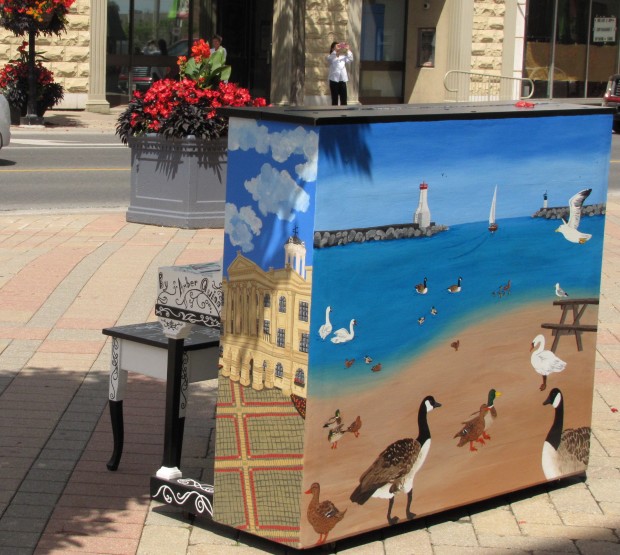 colour photograph of a street scene with the back and side of a piano in the foreground on a tiled sidewalk. On the side of the piano is a painting of Victoria Hall with a blue sky and on the back is a sunny harbour scene with a variety of ducks and swans in the foreground and the harbour entrance with lighthouse in the background. In front of the piano is a piano bench, at the near edge of the street is a large container of red flowers and across the street is a person taking a photograph.