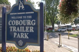 Une photo en couleurs avec une grande enseigne bleue indiquant le Cobourg Trailer Park et, de l’autre côté du chemin de terre, sous les arbres, on retrouve des remorques de villégiature et deux voitures.