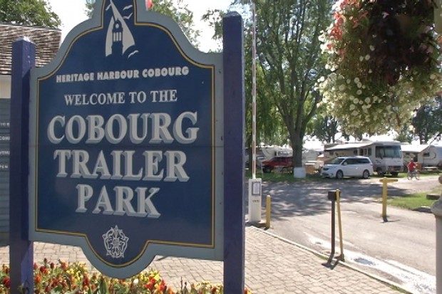 colour photograph with large blue sign identifying the Cobourg Trailer Park and across the dirt road under trees are some vacation trailer and two cars