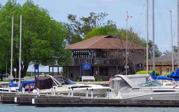a dark brown two-story clubhouse is surrounded by green trees with yachts moored in foreground