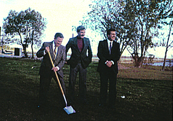 old colour photograph of three smiling men in suits posing on a patch of green grass with a shoreline in the background. The man to the left is inserting a shovel into the ground