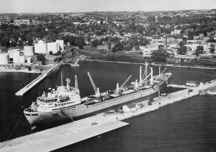 black and white photograph of a large freighter docked along a pier in the middle with a couple of cranes on board. A second pier leads back to oil tanks on shore to the left with piles of coal and a railway car along the shore between the two piers