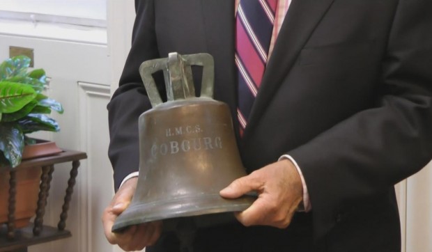 a colour photo of a man in suit and tie, shown only from the waist to the shoulders, holds a bronze ship's bell with HMCS COBOURG inscribed on it