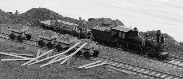 a black and white photograph of diorama reproducing a rare photograph. in the foreground planks of lumber lean against one of four ore cars on rails. Beyond is a steam engine with a tender full of lumber and a flatbed. Scattered on and around these are a dozen workers posing for the picture.