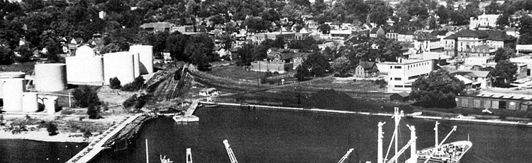 a black and white photo of an industrial harbourfront with white oil tanks to the left, railway lines and piles of coal in the middle and a boxcar to the right. In the right distance is Victoria Hall.