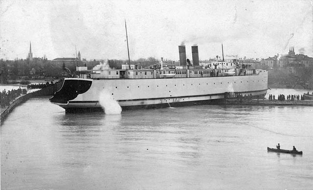 black and white picture of a large white ferry boat turning within tight confines between two wharfs. a small group of people watches on one wharf and two people in a canoe paddle in the foreground