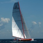 a colour photograph of a sleek red and white hulled sailboat on dark blue water with two white sails against a light blue sky. flying a Canadian flag