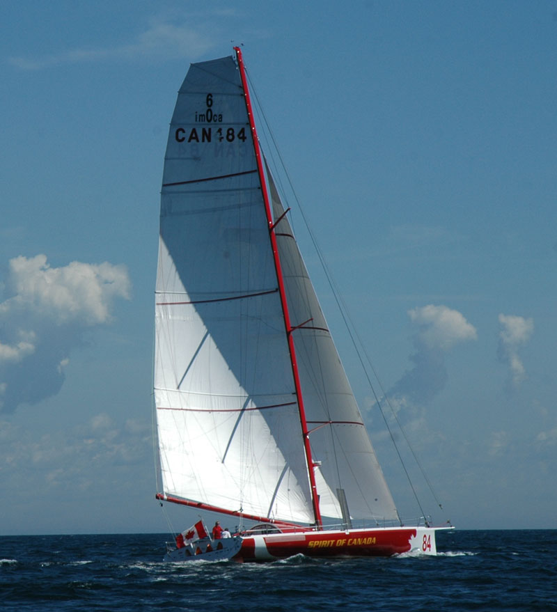 a colour photograph of a sleek red and white hulled sailboat on dark blue water with two white sails against a light blue sky. flying a Canadian flag