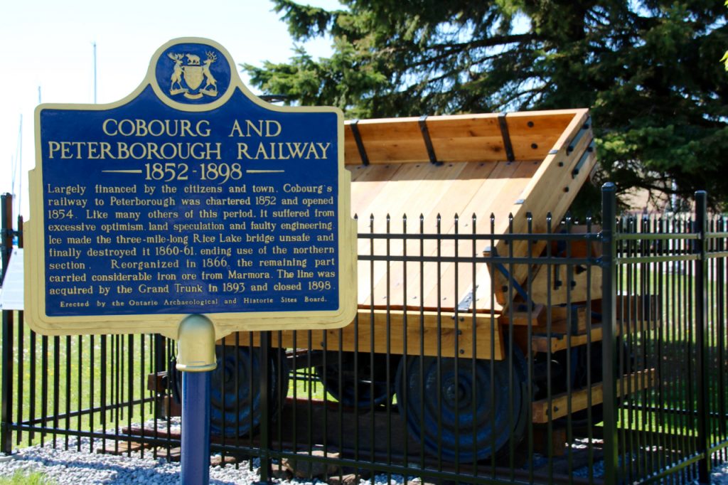 blue Province of Ontario history plaque tells the story of the Cobourg and Peterborough Railway in front of a full-size replica of an 1867 ore car, not far from where the original was built