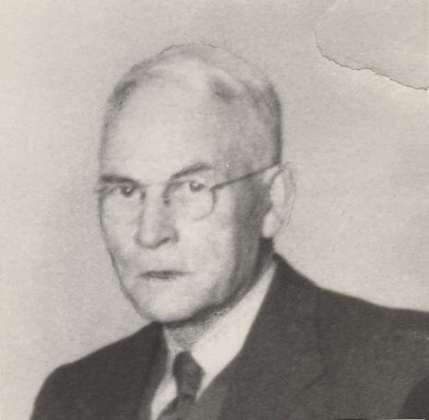 J.A. Campbell is staring at the camera and he looks severe. He is wearing glasses, a dark coloured jacket and vest, a lighter coloured shirt and tie.