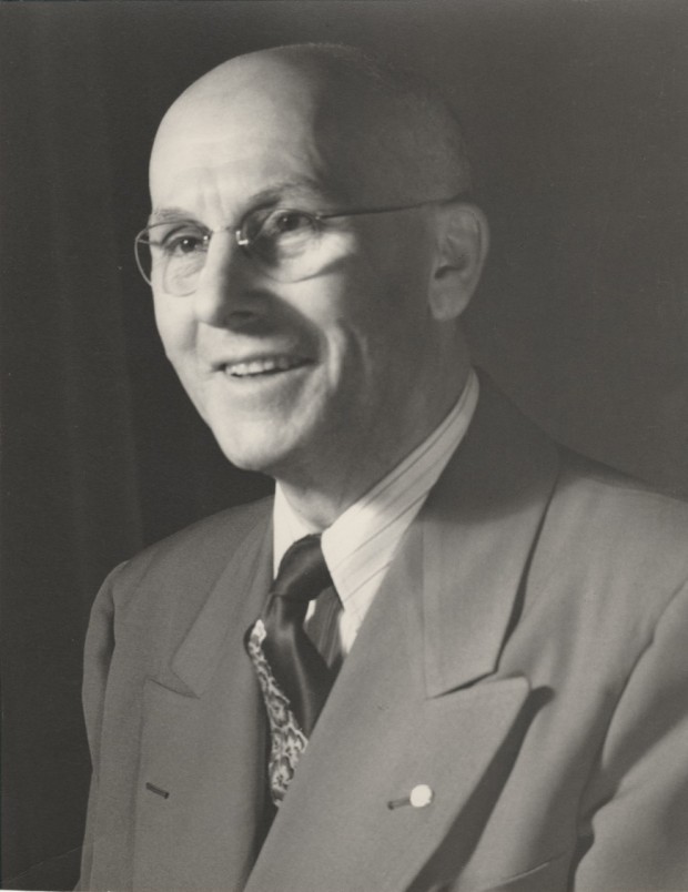 Henry Fishman is smiling in the photograph which appears to be taken in a dark room. He is wearing a light coloured jacket with a lighter coloured shirt and tie.