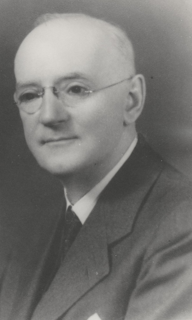 John Ridyard posing for a portrait photograph wearing glasses and a shirt, tie and jacket.