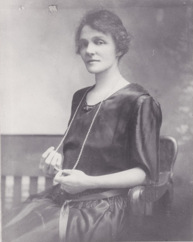 Violet Guymer as a young woman posing for a portrait. She is wearing a dark dress and has a string of pearls that extends to her lap.