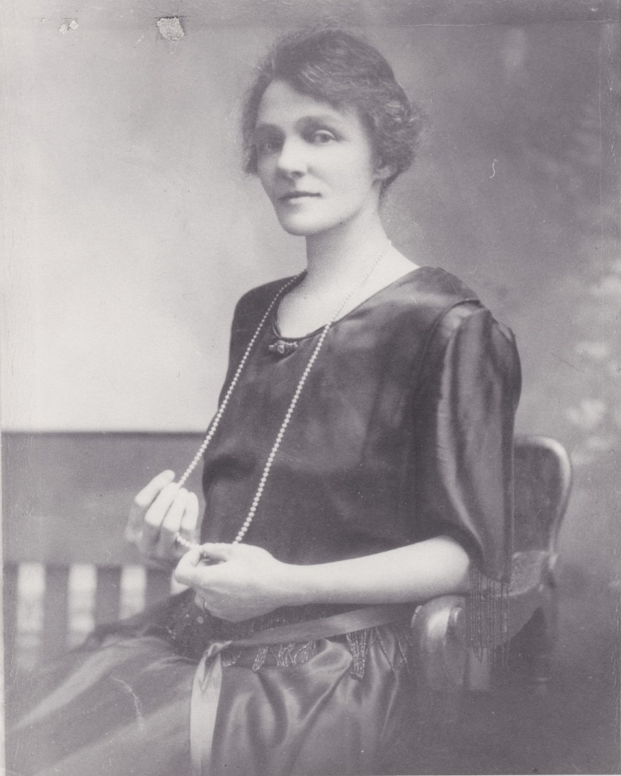 Violet Guymer as a young woman posing for a portrait. She is wearing a dark dress and has a string of pearls that extends to her lap.