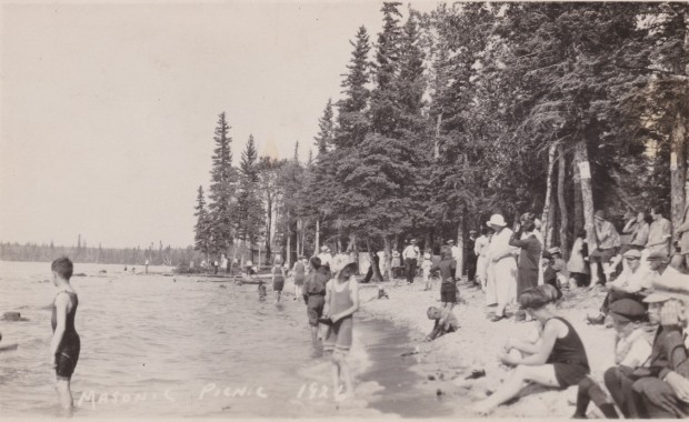 People are in their swimming outfits with a few of them wading into the water. There is little beach but lots of trees (primarily coniferous trees) in the background.