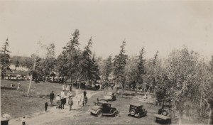 People are walking down a path going towards the water for the picnic. Cars are parked to the right and to the left there is open space. Numerous trees are in the background and through a space in the trees the lake is visible.