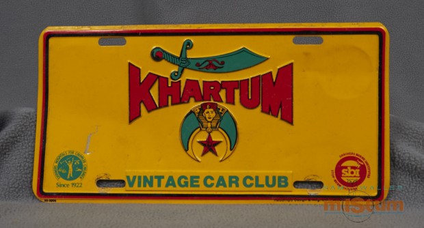 Golden yellow licence plate with red and green lettering and symbols of the Shriners club which consists of an upside down quarter moon filled in with green colouring, an Egyptian Pharaoh inside a pyramid at the top of the moon and a star hanging underneath the Pharaoh.