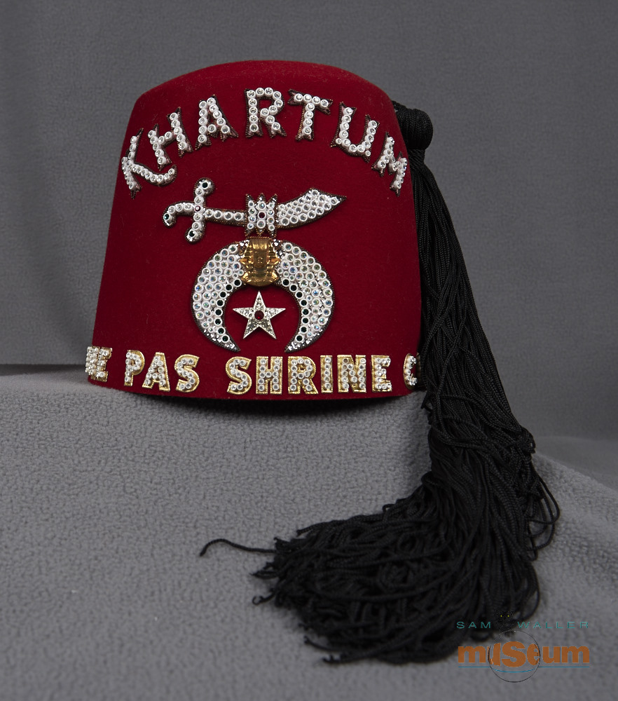 A red Shriners fez. On top there are four sets of air holes in the form of circles, and three rows of holes in each circle. The beaded lettering on the front reads "KHARTUM THE PAS SHRINE CLUB". There is also a symbol of an Arabic Sword and an Egyptian crest with a silver star below it. A long black tassel is attached from the top and hangs down the side. On the right side there is a silver broach with a studded Arabic sword and a crescent below with a maple leaf in the center.