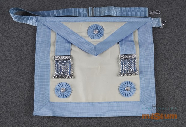 White apron with a blue border and strap that looks like an over-the-shoulder book bag. A flap, triangular in shape, hangs down 1/3 of the way centered by a silver mason badge with a blue floral design around it. The back is a piece of blue cotton fabric and the front is a white leather-material with blue bordering around the edges and front flap. Two blue cloth strips hang down with a metallic bar (silver) and 7 chains and small metal globes hanging from each bar on the bottom left and right corners are silver metal mason badges. In belt fastener is an S shaped silver snake. An adjustable belt with silver metal clasps is at the top under flap.