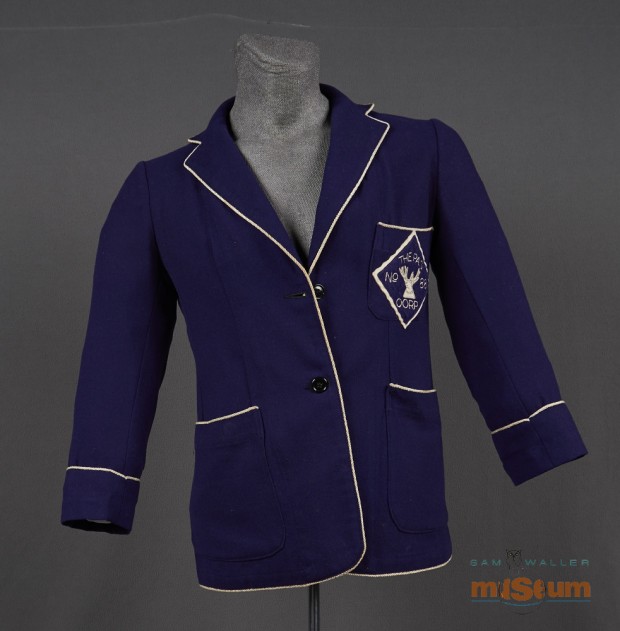 The jacket is made up of a purple textile and is single breasted. White piping is attached around all the edges except for the cuffs on the arms. The piping for the cuffs is located seven cm from the edge. There are three pockets on the jacket, one on either side at the bottom edge and a single pocket on the left breast. Attached to the breast pocket is The Pas Royal Purple crest.