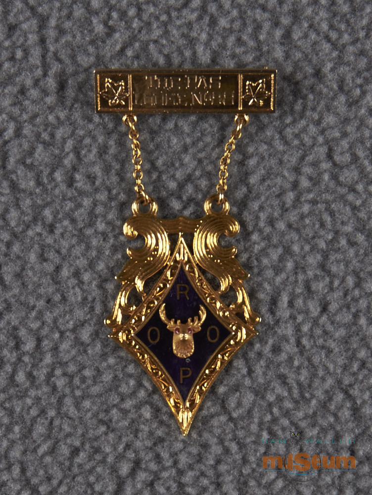 The main part of the pin is a three dimensional Royal Purple crest/logo with purple inlay. Of note, the eyes of the elk are inlaid with a red substance. There are ornate gold decorations above the crest/logo that lead to two chains. These chains hang from a pin that features two maple leaves and is inscribed 