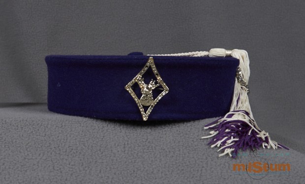 It is a bellboy style hat made from purple textile. A tassel attaches to a button on the top of the hat with a white cord. The tassel is made up of white and purple cords with a white cord tying them together near the top where the cords meet. A small chain of cut clear stones set in claws hold the tassel to the edge of the hat. On the front of the hat is a diamond shaped pin. The pin is an outline with an elk's bust in the centre. The diamond outline is studded with cut clear stones and the elk has cut orange stones for eyes.