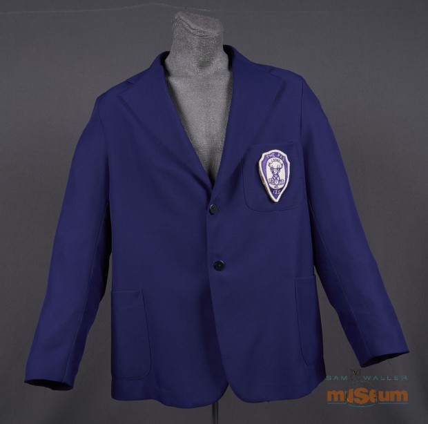 The jacket is single breasted and purple throughout. There is a partial inner lining of a slightly lighter purple. The jacket has two side pockets and one breast pocket on the left side. The Elks crest is sewn to this pocket. The crest reads The Pas/Lodge/B.P.O.E./No./135. The Pas and 135 are hand stitched to the crest while the rest is machine stitched.