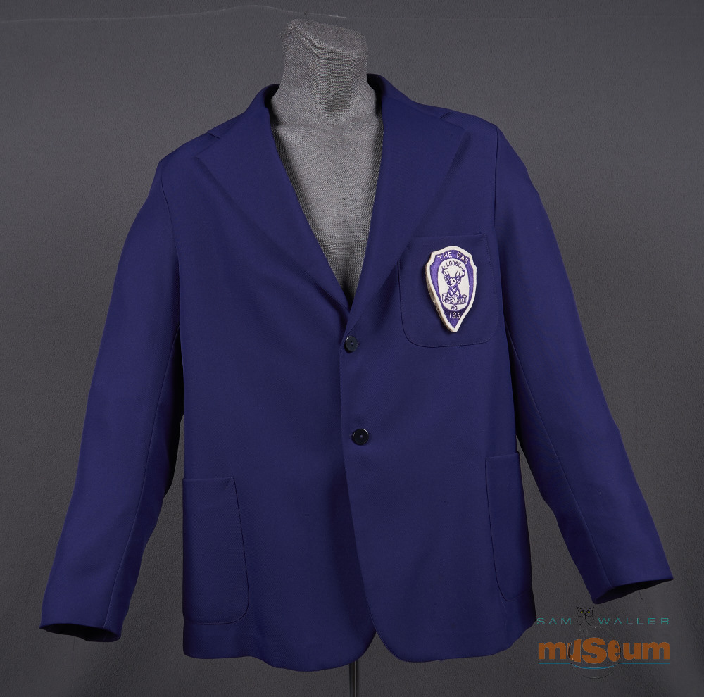 The jacket is single breasted and purple throughout. There is a partial inner lining of a slightly lighter purple. The jacket has two side pockets and one breast pocket on the left side. The Elks crest is sewn to this pocket. The crest reads "The Pas/Lodge/B.P.O.E./No./135". "The Pas" and "135" are hand stitched to the crest while the rest is machine stitched.
