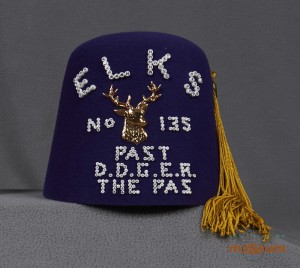 Un fez violet avec un gland doré et « Elks/No 135/Past/D.D.G.E.R./The Pas » inscrit dessus. Au centre de l’inscription se trouve un relief en métal représentant un élan.