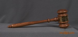 This gavel has a handle attached to a mallet style head, both made of wood and lightly stained with a varnish layer. Both parts are carved with simple lathe style designs. The middle of the mallet head is wrapped with a sheet of metal and the ends are fixed where the handle attaches to the mallet head. The metal sheet has the Lions emblem and the text: 