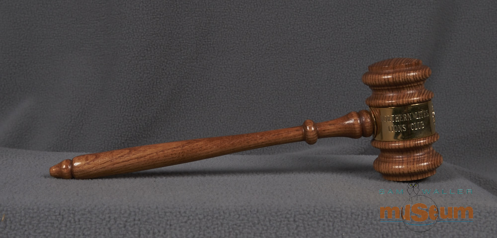 This gavel has a handle attached to a mallet style head, both made of wood and lightly stained with a varnish layer. Both parts are carved with simple lathe style designs. The middle of the mallet head is wrapped with a sheet of metal and the ends are fixed where the handle attaches to the mallet head. The metal sheet has the Lions emblem and the text: "Northern Lites/Lions Club" and "The Pas/Manitoba".