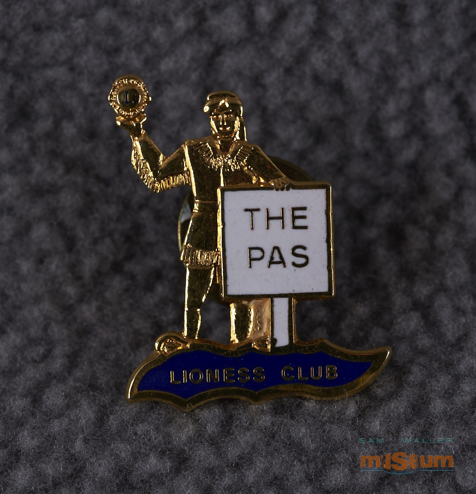 This pin is in the shape of the Trappers sign. The recto shows the man from the Trappers sign, in gold, holding the Lions emblem in his hand. There is a white sign in front of him with gold coloured lettering reading "The Pas". He and the sign are standing on an irregular shaped piece with a dark blue field.