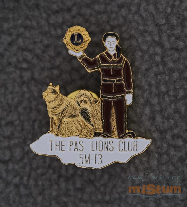 The gold pin highlights a trapper holding a Lions emblem in his hand. He has a white face and hands, a black hat and brown clothing. To his right is a sled dog, completely in gold. They are both standing on an irregular patch of white snow. The gold text “The Pas Lions Club/5M-13” appears in the in the snow patch. The text “Artiss-Regina/Taiwan” appears near the bottom of the textured gold back. This pin is cut to the shape of the Trapper, sled dog and snow patch and has a single butterfly clutch.