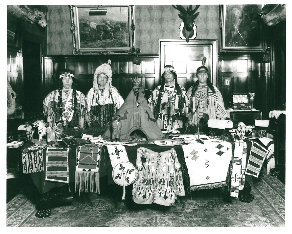 Interior shot at Beaulieu with 4 people dressed in Indigenous clothing behind a table with Indigenous objects; Southern Alberta Pioneer and Oldtimers Women's group in 1923