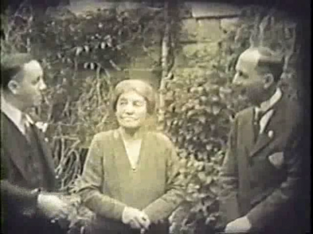 A period photograph of two well dressed men and a woman in a garden