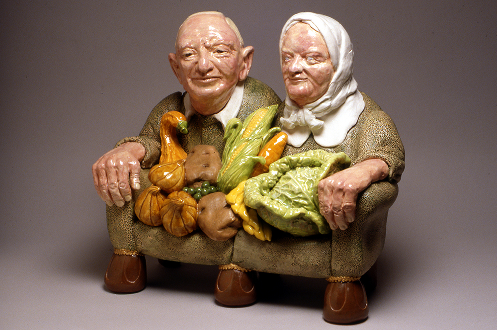 Sculpture en céramique d’un couple de personnes âgées enfoncé dans un canapé avec des légumes dans les mains