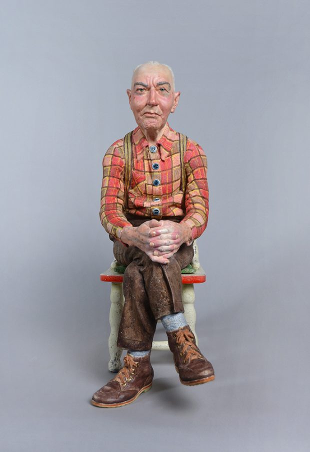 Sculpture of seated man in red and orange plaid shirt and brown pants.