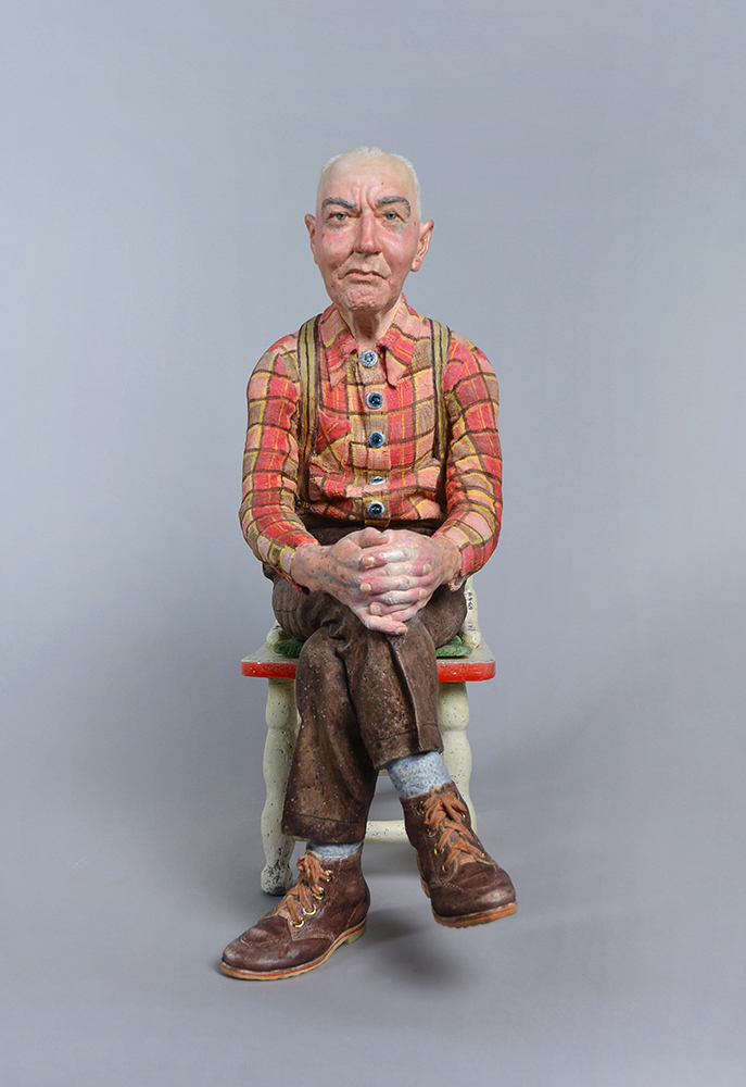 Sculpture représentant un homme assis qui porte une chemise à carreaux rouge et orange et un pantalon brun.