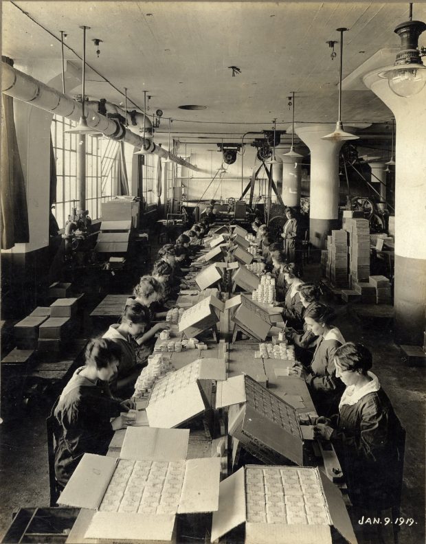 Two rows of women work at a long table in a warehouse in a black and white photo. Boxes and factory equipment in the background.
