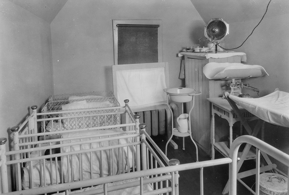 Cinq lits de bébé alignés le long du mur dans une petite pièce avec balance, cuvette et poste à langer situé du côté opposé dans une photo en noir et blanc.