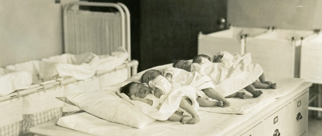 Sleeping newborn babies lined up in a row on a table with cribs in the background in a black and white photo.