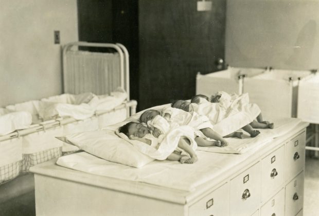 Sleeping newborn babies lined up in a row on a table with cribs in the background in a black and white photo.