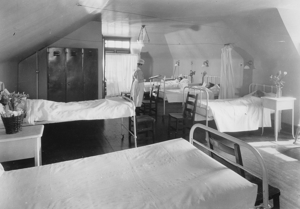 An old black and white photo of a room in a hospital showing five beds, four with patients. A nurse stands at the end of one reading a chart.