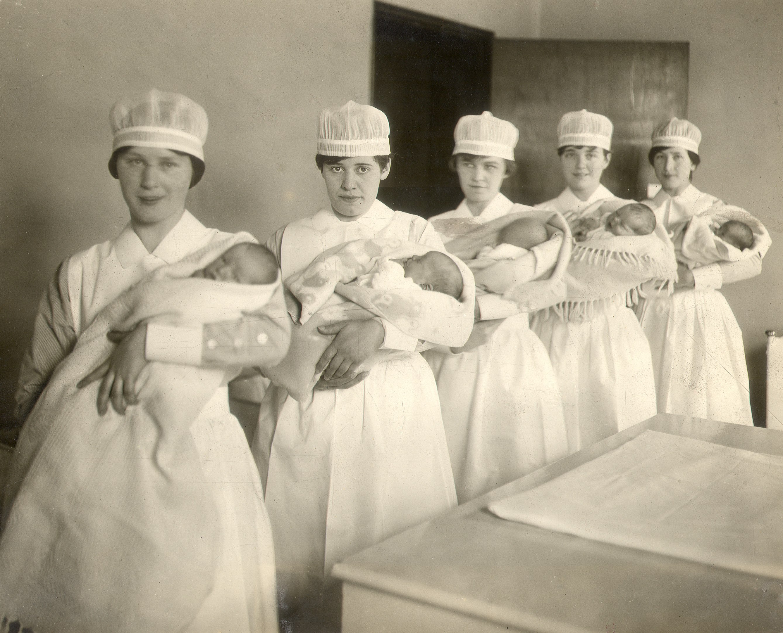 Five nurses in uniform hold swaddled babies beside a large table in a black and white photo.