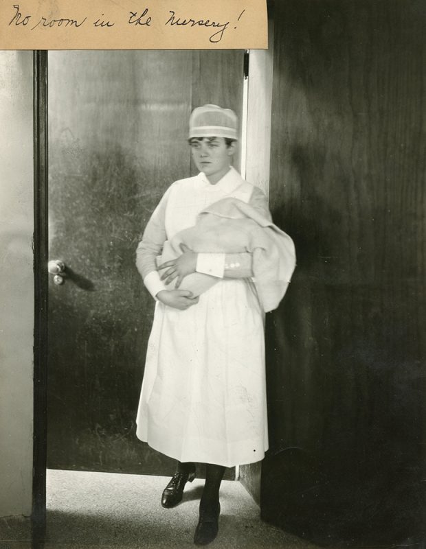 A nurse in uniform holds a newborn wrapped in a blanket as she exits a room in a black and white photo.