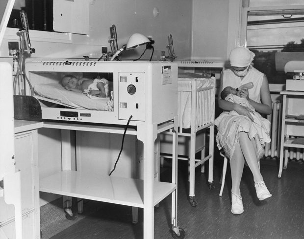 A nurse wearing a mask feeds a baby sitting beside a crib in a black and white photo. A second baby is in an incubator in the left foreground.