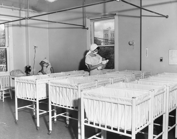 Two nurses wearing masks in a nursery full of white baby cribs in a black and white photo. Each holds a newborn.